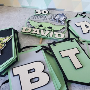 May include: A green and black banner with the letters "B", "T", and "A" in white. The banner is decorated with a green and black character from the Star Wars franchise. The banner also includes a cake topper with a character from the Star Wars franchise and the text "30 DAVID".