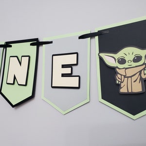 May include: A banner with the word "ONE" and a Grogu character design. The banner features gray, black, and light green pennants with black ribbon for hanging. The Grogu image is on a black pennant.