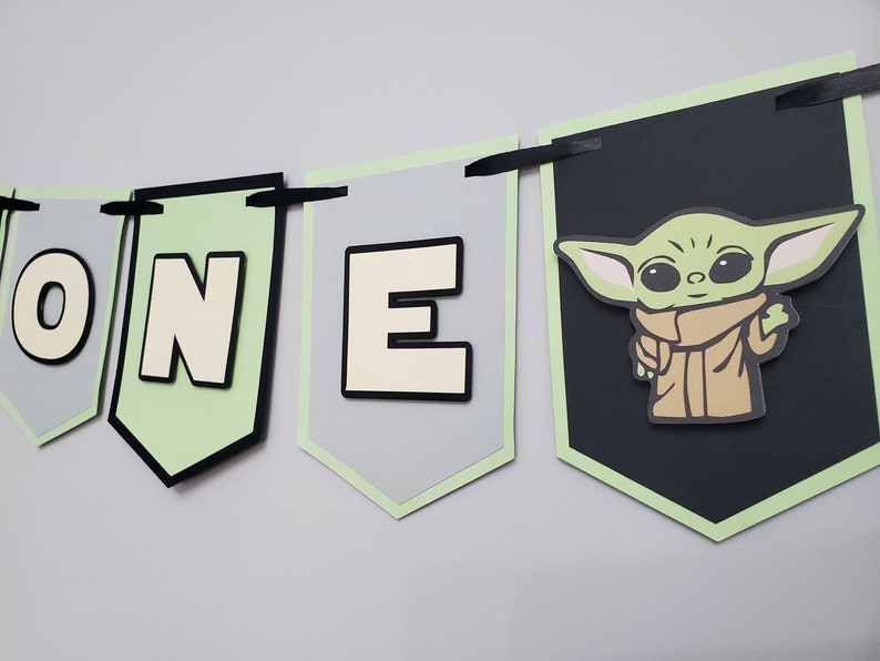 May include: A banner with the word "ONE" and a Grogu character design. The banner features gray, black, and light green pennants with black ribbon for hanging. The Grogu image is on a black pennant.