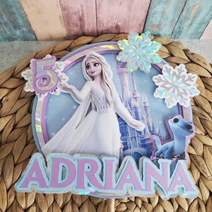 Könnte beinhalten: Ein individueller Tortenaufsatz mit der Figur Elsa aus dem Film Frozen. Der Aufsatz enthält den Namen "ADRIANA" in Lavendel und Hellblau, die Zahl "5" und Schneeflocken-Akzente. Der Hintergrund zeigt ein Schloss und eine blaue Kreatur.
