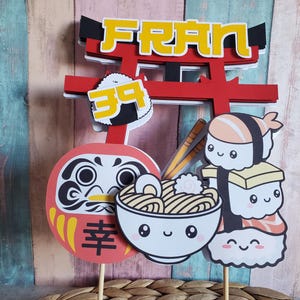 May include: A vibrant Japanese-themed cake topper with a red torii gate, the name "FRAN" in yellow, and the number "39". It features illustrations of sushi, ramen, and a Daruma doll. The background is a mix of wood panels.