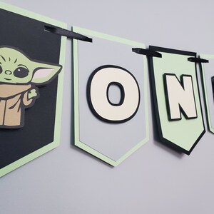 May include: A banner with a green and black color scheme, featuring the character Grogu from The Mandalorian. The banner spells out "ONE" with a light green border and black ribbon for hanging. The Grogu image is on a black banner.