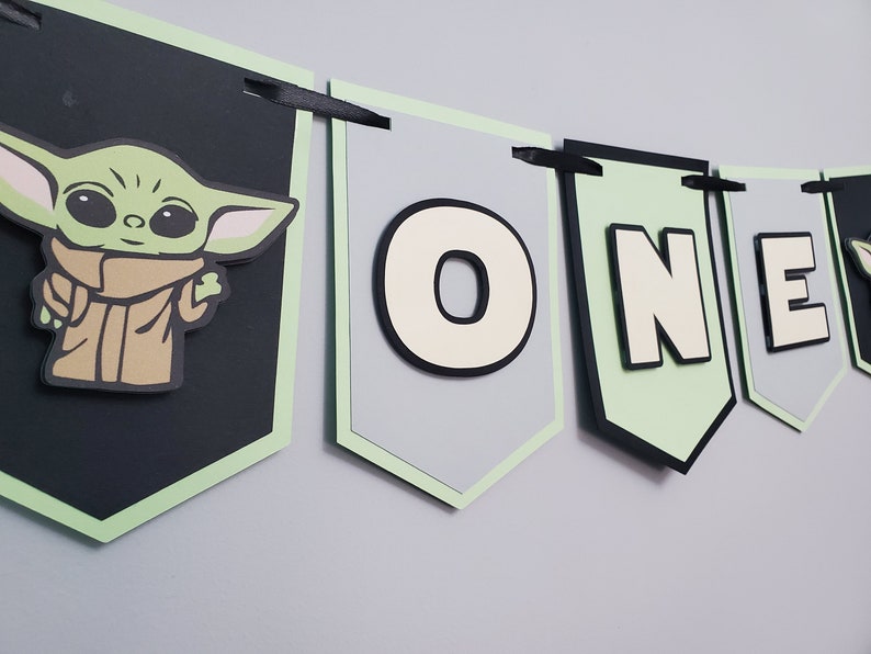 May include: A banner with a green and black color scheme, featuring the character Grogu from The Mandalorian. The banner spells out "ONE" with a light green border and black ribbon for hanging. The Grogu image is on a black banner.