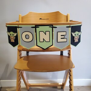 May include: Wooden high chair with a banner that says "ONE". The banner has green, gray, and black pennants with a cartoon character and the letters "O", "N", and "E". The high chair is made of light-colored wood.