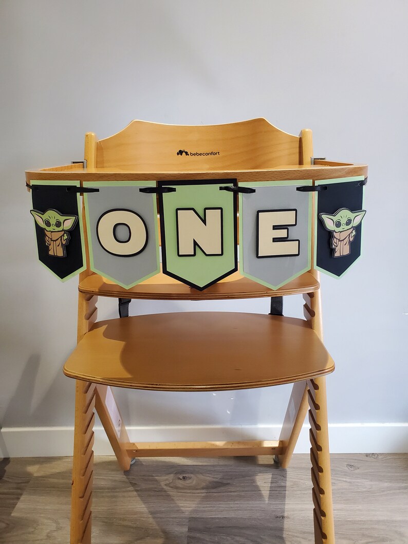 May include: Wooden high chair with a banner that says "ONE". The banner has green, gray, and black pennants with a cartoon character and the letters "O", "N", and "E". The high chair is made of light-colored wood.
