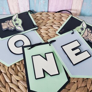 May include: A banner set with the word "ONE" and two tags featuring a cartoon character. The letters are white with black outlines, set against light green and gray backgrounds. The character tags are black with a light green border.