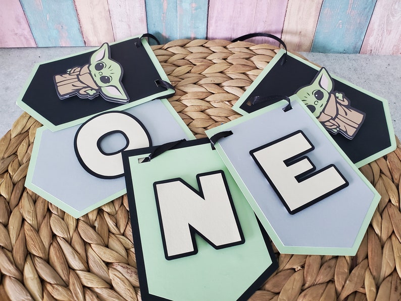 May include: A banner set with the word "ONE" and two tags featuring a cartoon character. The letters are white with black outlines, set against light green and gray backgrounds. The character tags are black with a light green border.