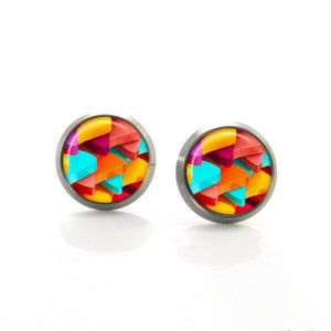 Könnte beinhalten: Ein Paar runde Ohrstecker mit einem farbenfrohen geometrischen Design. Die Ohrringe haben eine Edelstahlfassung und ein lebendiges Muster aus orangefarbenen, gelben, roten, rosa und türkisfarbenen Formen. Die Ohrringe werden auf einer weißen Oberfläche präsentiert.