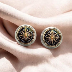 Puede incluir: Dos gemelos de plata con diseño de brújula. La cara de la brújula es negra con detalles dorados y presenta las direcciones cardinales.