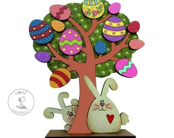 May include: A wooden Easter tree decoration with a brown trunk and green leaves. The tree is decorated with colourful Easter eggs and two white bunnies are peeking out from behind the trunk. The bunnies have red hearts on their chests. The base of the tree is a brown wooden platform. The image includes the text "Hobby Market".
