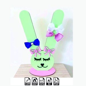 May include: A green and pink bunny-shaped hair bow holder with a pink base. The holder has two ears and a face with eyelashes and a heart-shaped nose. There are three bows on the ears, one blue, one pink, and one white.