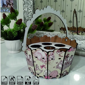 Könnte beinhalten: Ein weißer, hölzerner Eierhalter in Form eines Korbes mit Henkel. Der Korb hat ein florales Design mit rosa und braunen Akzenten. Der Korb hat sechs Fächer für Eier.