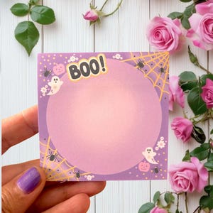 May include: A square, lavender-colored Halloween-themed sticky note with the word "BOO!" in yellow. The note features a spider web design, spiders, ghosts, and pumpkins. The note is held in a hand, with pink roses in the background.