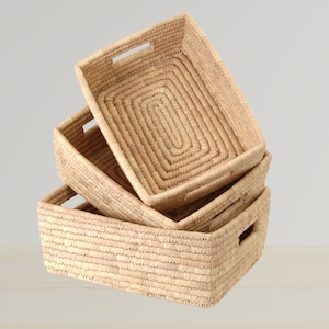 Puede incluir: Tres cestas tejidas de materiales naturales, apiladas una encima de la otra. Las cestas son rectangulares y de color marrón claro.