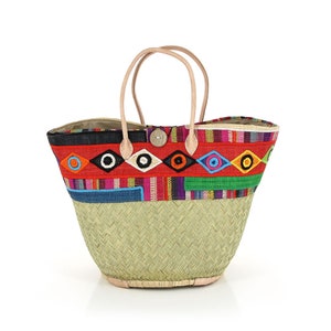 Könnte beinhalten: Eine gewebte Stroh-Tragetasche mit einem farbenfrohen Stickdesign. Die Tasche hat ein rotes Band mit gestickten Augenformen, einen Holzknopf und Ledergriffe. Die Basis der Tasche ist hellgrün.