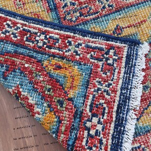 May include: Close-up of a colorful rug with a blue, red, and yellow pattern. The rug has a fringe border and is folded over on a wooden surface.