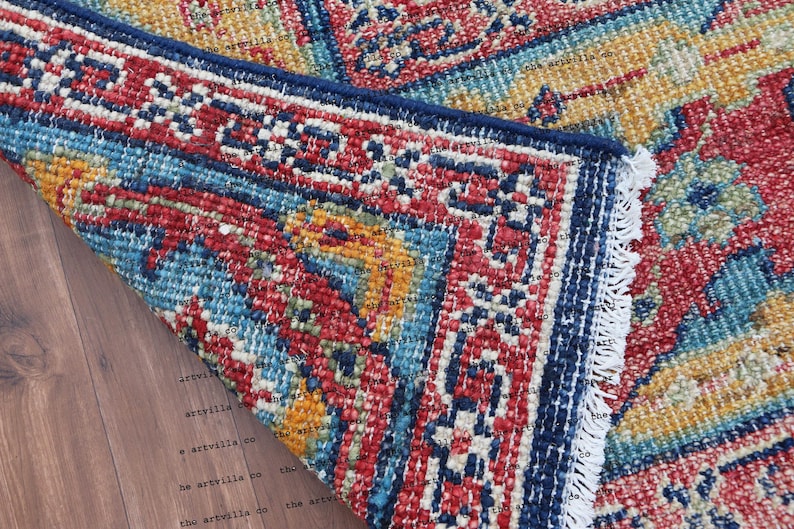 May include: Close-up of a colorful rug with a blue, red, and yellow pattern. The rug has a fringe border and is folded over on a wooden surface.