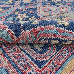 May include: A close-up of a blue, red, and yellow patterned rug. The rug has a traditional design with intricate details. The rug is folded in half, showing the texture and weave of the fabric.