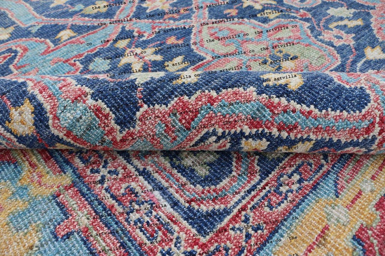 May include: A close-up of a blue, red, and yellow patterned rug. The rug has a traditional design with intricate details. The rug is folded in half, showing the texture and weave of the fabric.