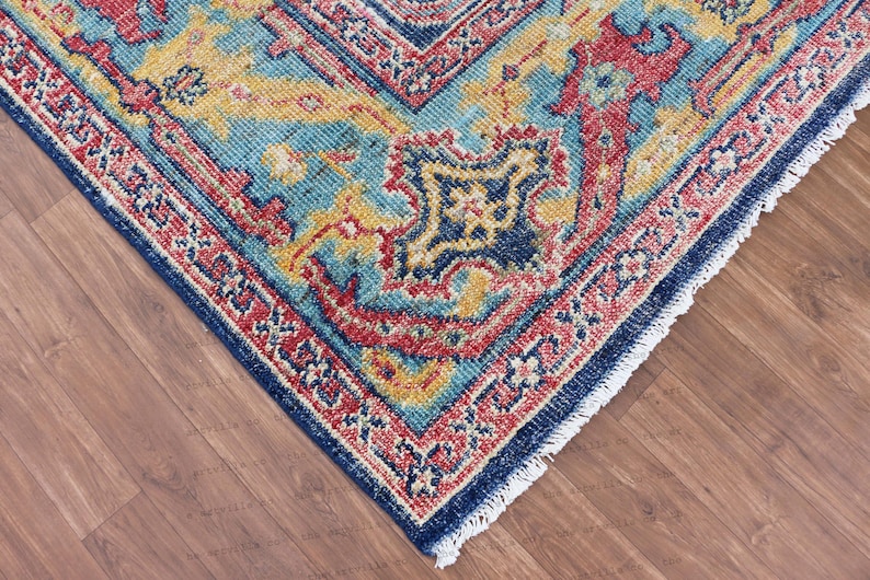 May include: Close-up of a blue, red, and yellow patterned rug with a fringed edge. The rug has a traditional design with intricate details.