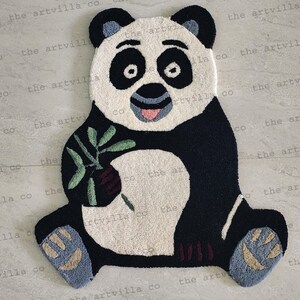 Puede incluir: Una alfombra de panda blanco y negro con una hoja verde en su pata. La alfombra está hecha de un material suave y mullido y tiene una expresión juguetona.