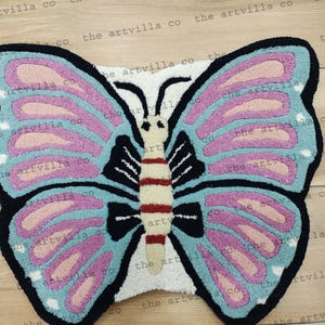 May include: A blue and pink butterfly rug with a white center. The butterfly has a black outline and a striped body.