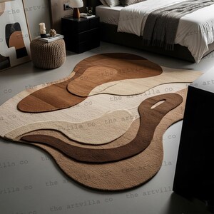Puede incluir: Una alfombra abstracta en tonos marrones y beige, con un diseño único y en capas. La forma orgánica de la alfombra está sobre un suelo gris. El texto "the artvilla co" está impreso en la alfombra. Dimensiones: aprox. 200 cm x 300 cm.