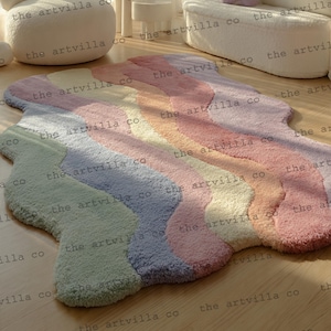 Puede incluir: Una alfombra ondulada multicolor en tonos pastel de rosa, naranja, amarillo, verde y azul. La alfombra tiene una textura suave y afelpada y está colocada sobre un suelo de madera. La forma única y los colores vibrantes de la alfombra la convierten en un acento decorativo.
