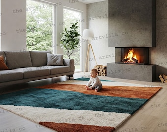 May include: A large, rectangular area rug with a modern design. The rug features bands of color: rust, teal, white, and brown. The rug is placed in a living room with a gray sofa, fireplace, and large windows.
