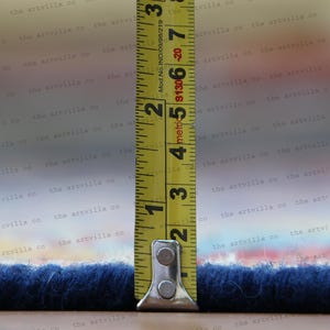 May include: A yellow measuring tape with black markings is extended to measure the width of a blue rug. The tape reads "metric 5 4 3 2 1" and "Mod. No. IND/09/98/219 $1306-20 7 3".