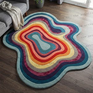 Puede incluir: Una alfombra abstracta multicolor con una forma irregular. La alfombra presenta bandas concéntricas de color, incluyendo rojo, naranja, amarillo, verde, azul y morado. La alfombra está colocada sobre un suelo de madera.