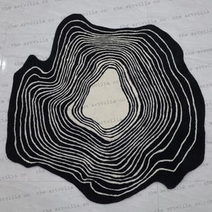 May include: Black and white abstract rug with a circular design resembling tree rings. The rug has a unique, organic shape.