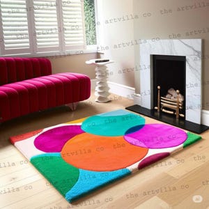 Peut inclure: Un tapis rectangulaire avec un motif abstrait coloré. Il présente de grands cercles superposés en bleu, vert, orange, rose et violet sur un fond blanc. Le tapis est posé sur un sol en bois.