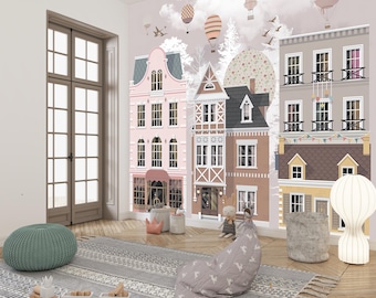 May include: A whimsical nursery scene featuring a wall mural of colorful townhouses with hot air balloons and birds. The room includes a green ottoman, a patterned rug, a beanbag chair, and a decorative lamp. Wooden toy train set is on the floor.