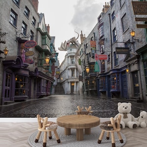 May include: A wallpaper mural depicting a cobblestone street lined with shops and buildings, reminiscent of a fantasy setting. A dragon statue is perched atop a building. The scene includes a round wooden table, chairs, and teddy bears.