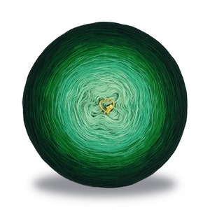 Può includere: Un gomitolo di lana verde con un gradiente dal verde scuro al verde chiaro. La lana è avvolta in un cerchio e ha un piccolo ciondolo a forma di cuore dorato al centro.