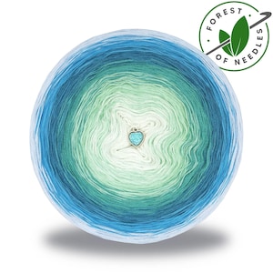 Può includere: Un gomitolo di filato con una sfumatura di colori, che vanno dal blu chiaro al blu scuro, con verde e bianco al centro. È attaccato un piccolo ciondolo a forma di cuore. L'immagine include un logo che dice "FOREST OF NEEDLES".