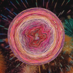 Puede incluir: Una vibrante madeja de hilo multicolor con tonos de rosa, rojo, naranja y morado, acentuada con hilos dorados. El hilo está dispuesto en círculos concéntricos, creando un efecto de espiral. Un pequeño objeto decorativo se encuentra en el centro. El fondo presenta un espectáculo de fuegos artificiales.