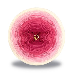 May include: A skein of yarn in a gradient of pink, white, and cream colours. The yarn is wound into a ball shape and has a small gold heart charm in the centre.