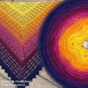 May include: A handmade crocheted shawl in a gradient of colours, shifting from yellow to orange, pink, and deep purple. The shawl features an open, lacy pattern. A ball of yarn with matching colours is next to the shawl. The text "Jamie D. Huskyberry Freya (Huskyberry)" is visible.