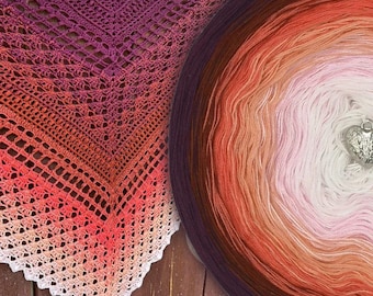 May include: A triangular crochet shawl featuring a gradient color transition from deep purple to bright orange, pink, and white. Beside it sits a matching circular yarn cake with the same color progression and a small silver heart-shaped charm.