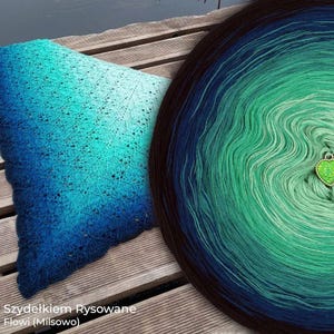 May include: A triangular crochet shawl featuring a blue to turquoise gradient and a circular yarn cake with a green to dark blue ombre transition, accented by a small heart-shaped charm. The text "Szydełkiem Rysowane Flowi (Milsowo)" is visible.