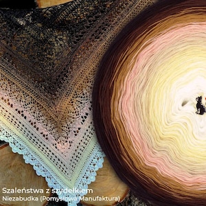 May include: A crocheted shawl with a gradient color scheme, transitioning from dark brown to light pink and yellow. The shawl has a delicate, lacy pattern. Next to it is a large ball of yarn with a similar color gradient. The text "Szaleństwa z szydełkiem Niezabudka (Pomysłowa Manufaktura)" is visible.