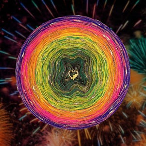 Puede incluir: Una bola de hilo multicolor con un dije en forma de corazón en el centro. El hilo muestra un espectro de colores, incluyendo rosa, naranja, amarillo, verde y azul, con hilos metálicos dorados. El fondo presenta una imagen borrosa de fuegos artificiales.