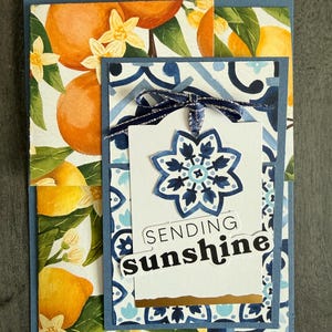 May include: Greeting card with a watercolor design of oranges and lemons. The card features a blue and white patterned background and a tag that says "SENDING sunshine". A blue ribbon and a decorative flower are attached to the tag.