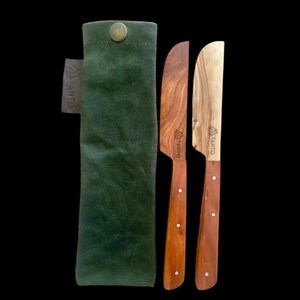 Könnte beinhalten: Ein Set aus zwei Buttermessern aus Holz mit Griffen aus Olivenholz und einer grünen Stoffhülle. Die Messer sind mit dem Wort "Taito" graviert.