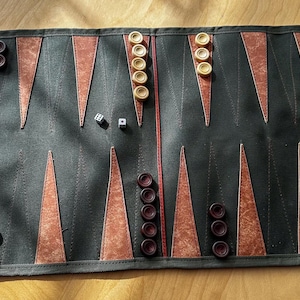 May include: A rolled-up backgammon board in dark green with brown triangular markings. The board is set with brown and cream-colored game pieces and two dice. A zip and a red strap are also visible.