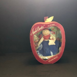 May include: A vintage Apple Cheeks collectible doll, Miss Wilma Wealthy, displayed in a red apple-shaped box. The doll wears a red top, blue polka-dot dress, and holds a smaller doll. The box has a clear window and the words "COLLECTIBLE" and "Apple Cheeks".