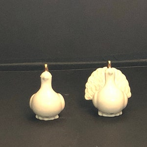 May include: Two ceramic turkey figurines. One turkey is facing forward, and the other has its tail feathers fanned out. Both are off-white with gold accents on their heads. The figurines are on a dark surface.
