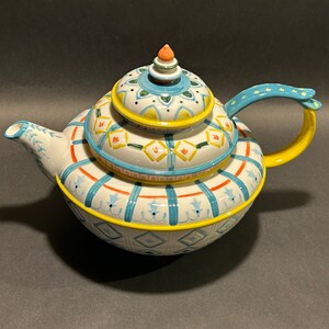 Puede incluir: Tetera decorativa de cer&aacute;mica con tapa, con base blanca adornada con dise&ntilde;os azules, naranjas y amarillos. El asa y los detalles son amarillos y azules. La tetera tiene un pico y un peque&ntilde;o adorno en la tapa.